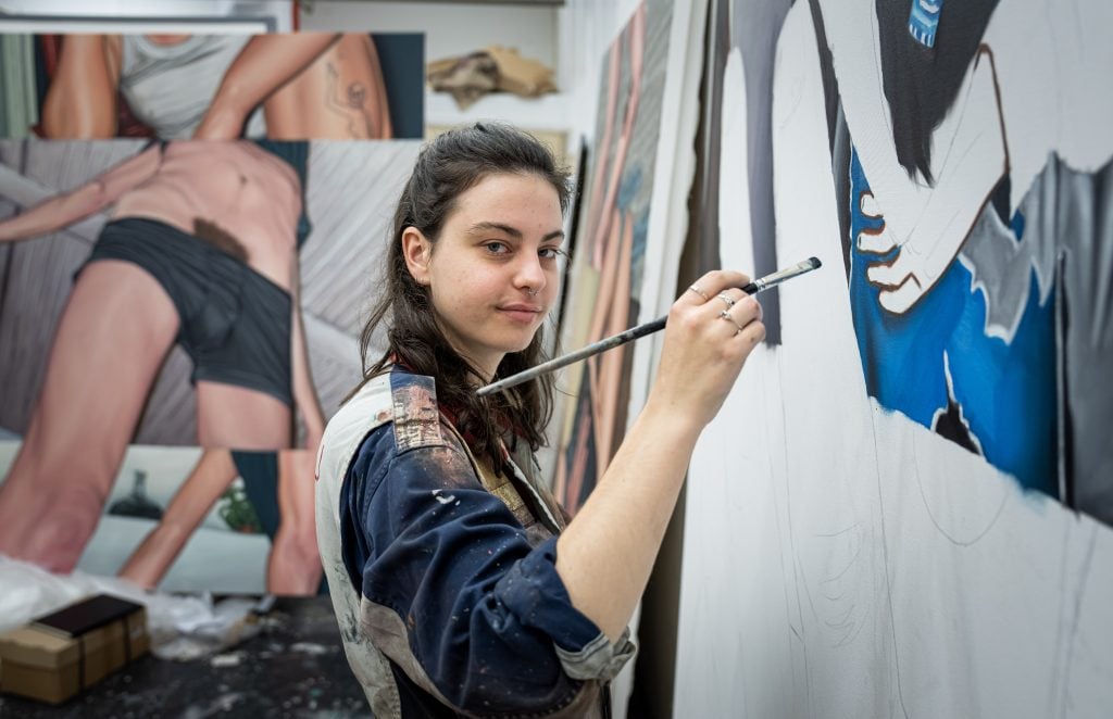 Half portrait of the artist Bobbi Essers holding a paintbrush to an unfinished painting surrounded by other paintings in their studio.