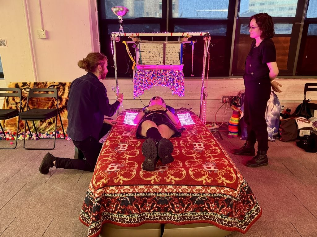 One of the late Bobby Anspach's <em>Continuous Eye Contact</em> works at Spring Break Art Show New York 2024. A person is lying down on a hospital bed draped in an oriental rug, beneath pom poms illuminated by LED lights. Two people on either side are helping operate the artwork. 