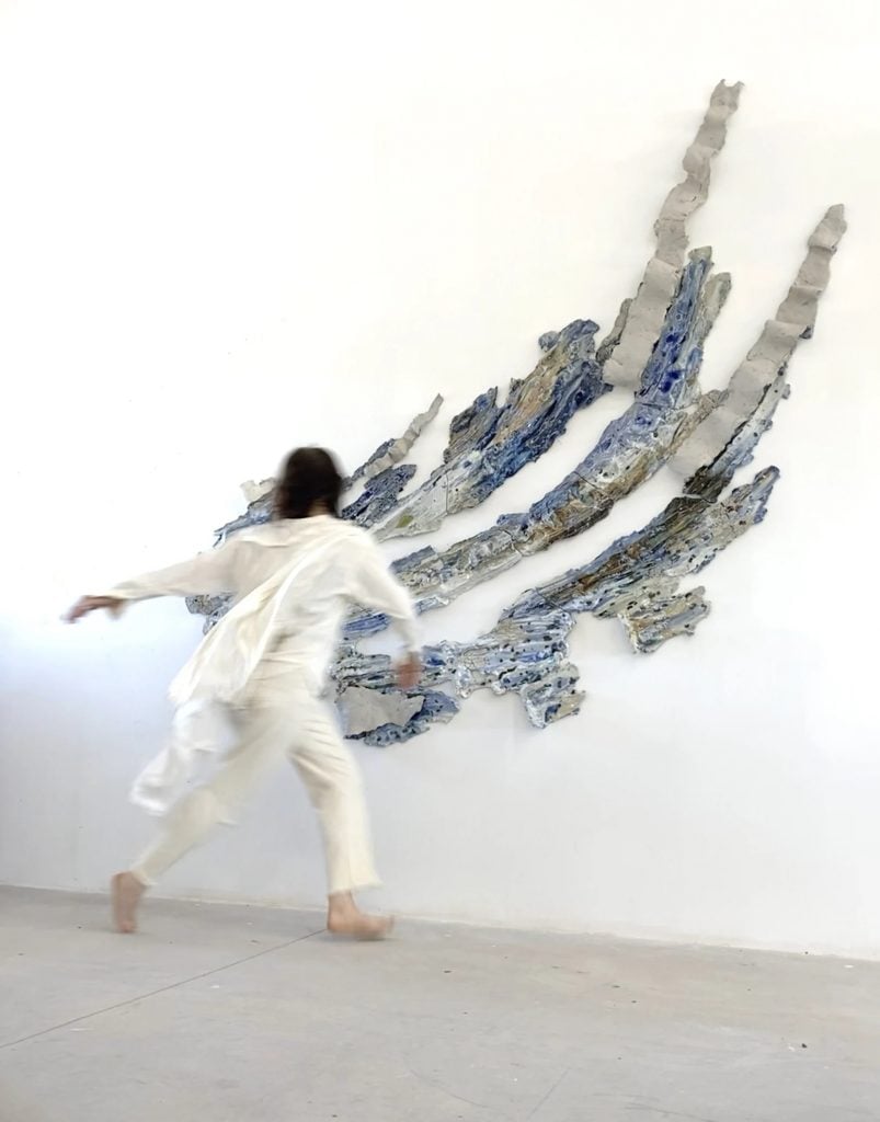 Brie Ruais with her clay sculpture "Traveling with the Wind, East, 130lbs" (2024). The piece is thin pieces of clay spread into long swooping forms and painted in shades of gray and brown and are inspired by gusts of wind. The artist is blurry, captured in motion with her back to the camera, wearing the flowing white garment she used while making the work.