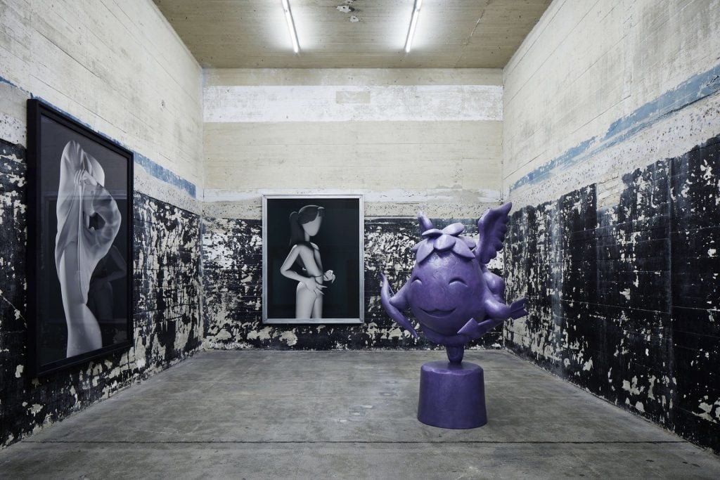 Inside a bunker room with two CGI flat images on the far and left wall, and a purple amorphic sculpture in the center.