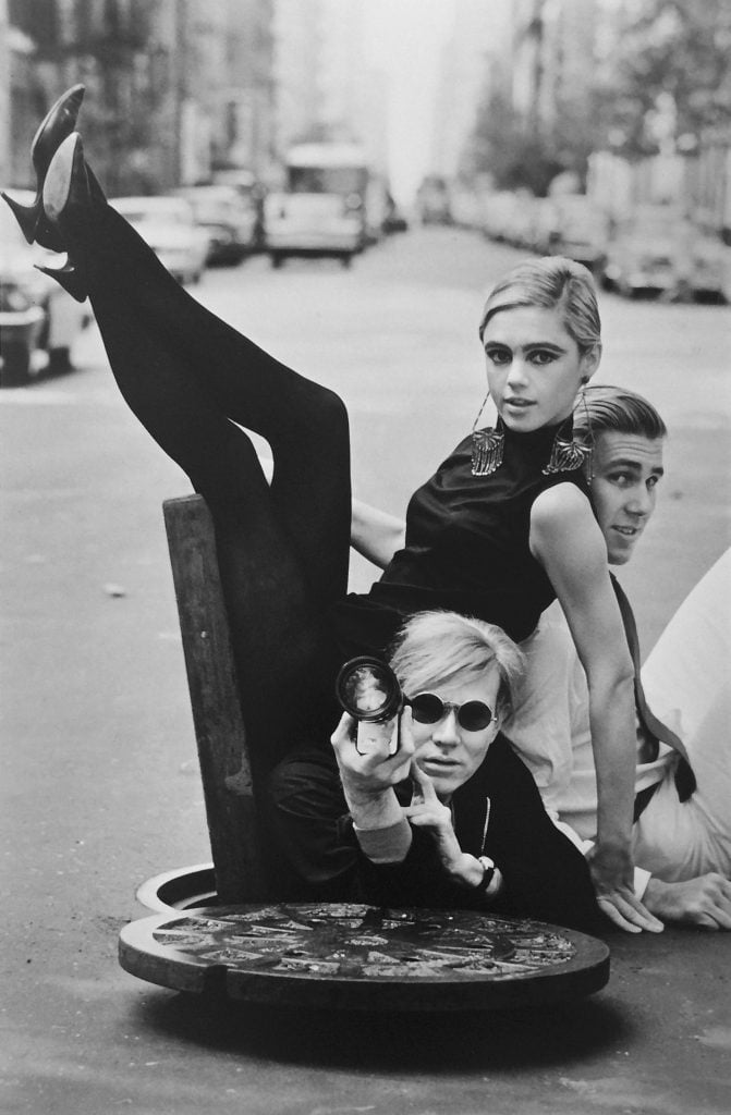 Black and white photo of Edie Sedwick, Andy Warhol, and Chuck Wein in mod clothing coming out of a manhole in the middle of an NYC street.