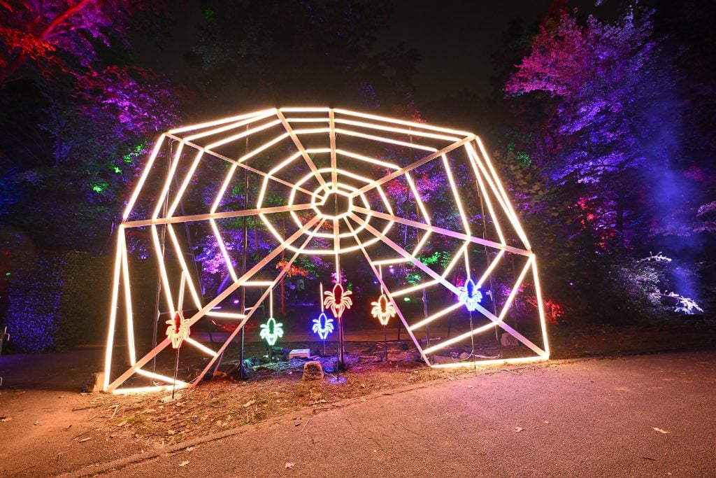 A photograph of a large, illuminated spider web with small illuminated spiders dangling from it.