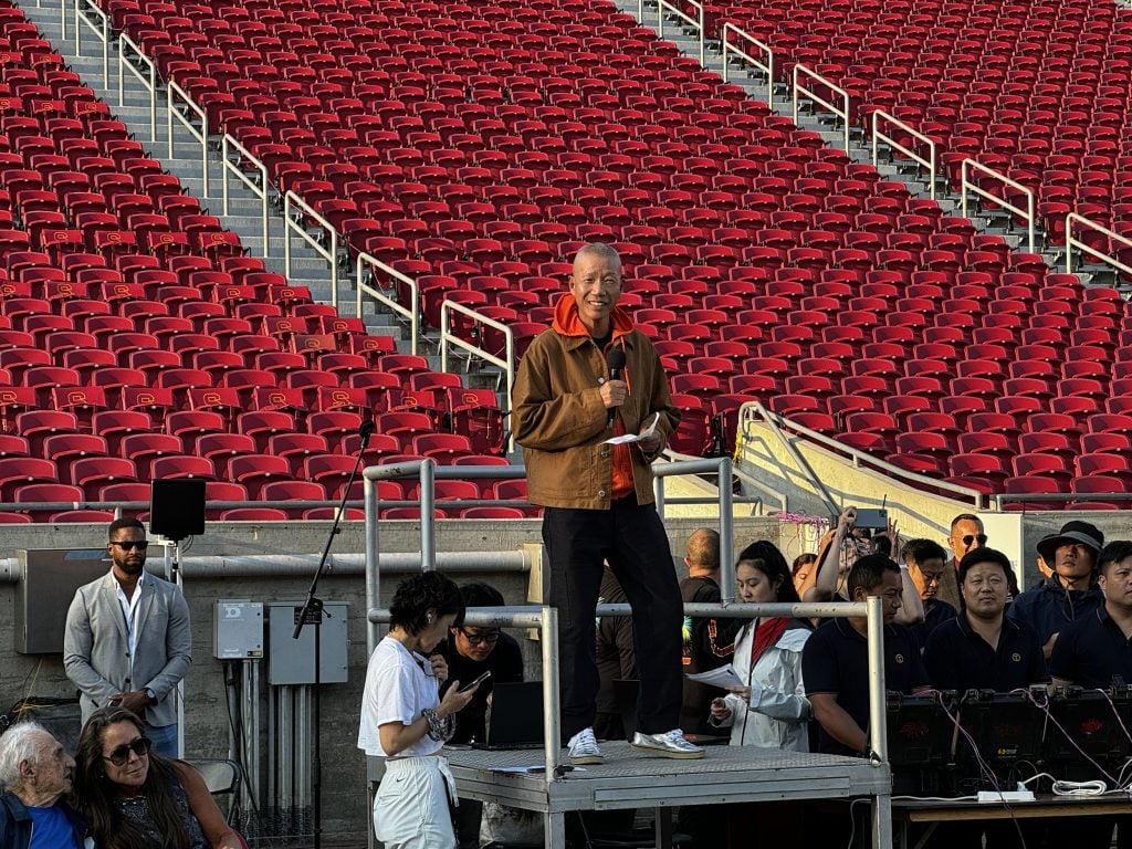 A man, Chinese artist Cai Guo-Qiang, standing on a platform in a brown jacket and orange hoodie, surrounded by a crew