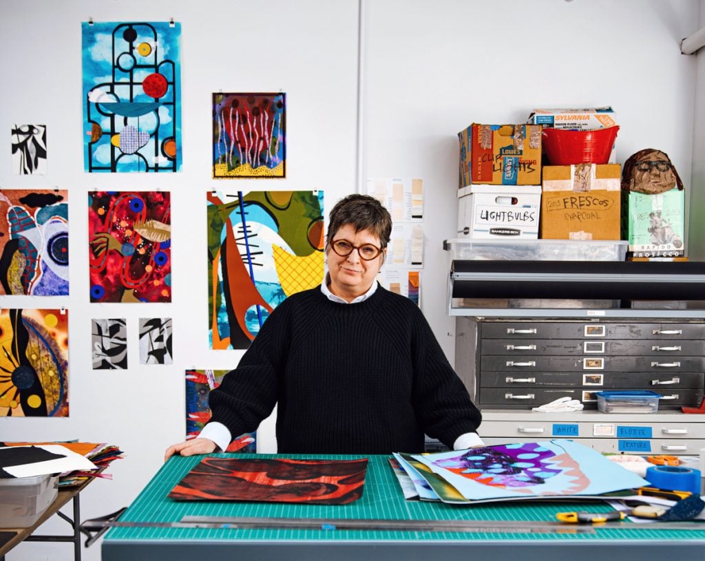 Portrait of artist Carrie Moyer at her studio work table wearing a black pullover over a white shirt, with small-scale artworks on the wall behind her, a document flat cabinet to her left, and a series of small works on paper on a cutting mat in front of her.