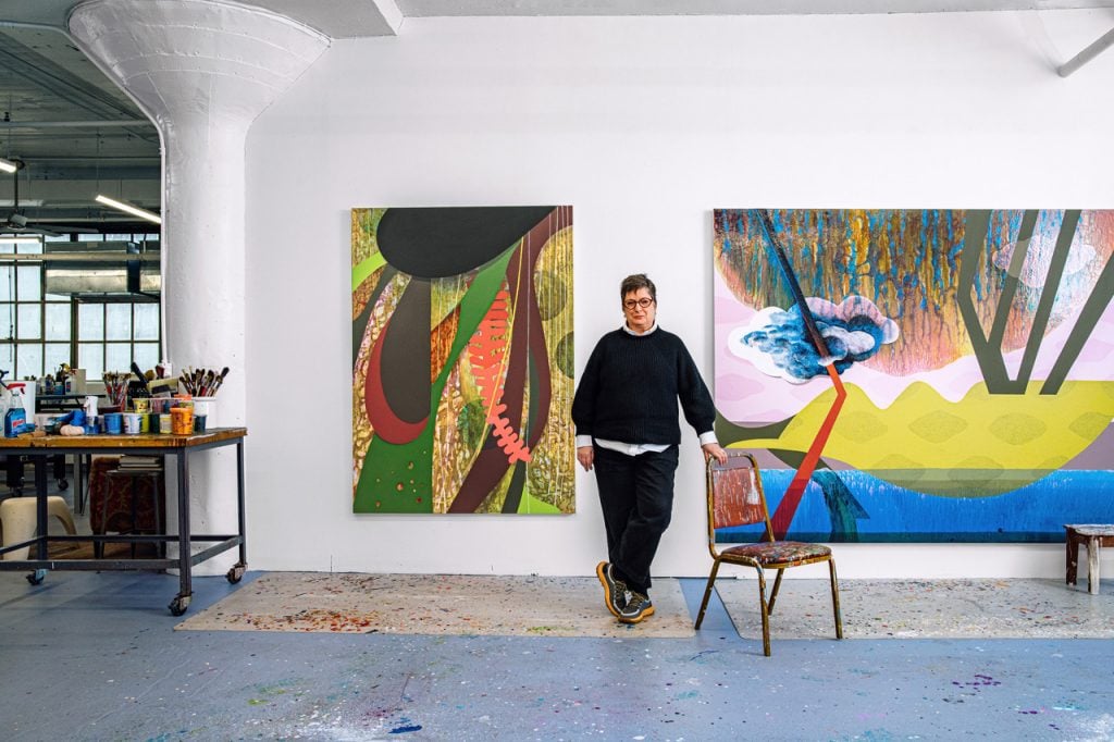 Portrait of Carrie Moyer in her studio standing in front of two large-scale paintings on the wall leaning on a red chair covered in paint splatters, wearing a black pullover and black pants. To the left a large industrial support column and a wall of windows can be seen.