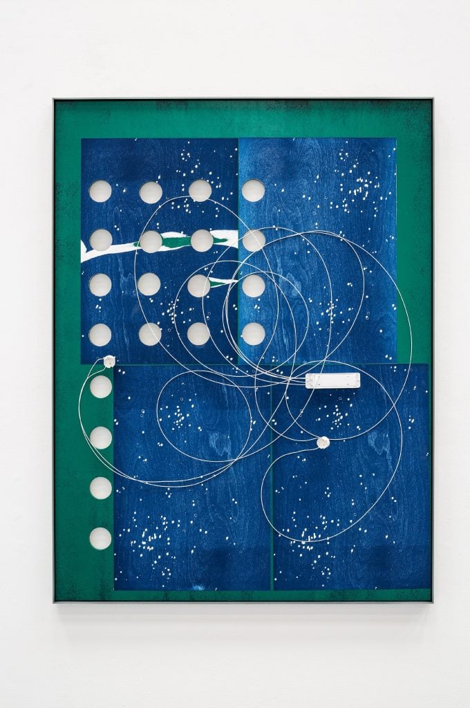 An abstract multimedia work in blue and green with a series of holes drilled through showing the white wall and looking like a moon calendar, with celestial orbits etched across the surface. Part of the space-inspired artworks roundup.