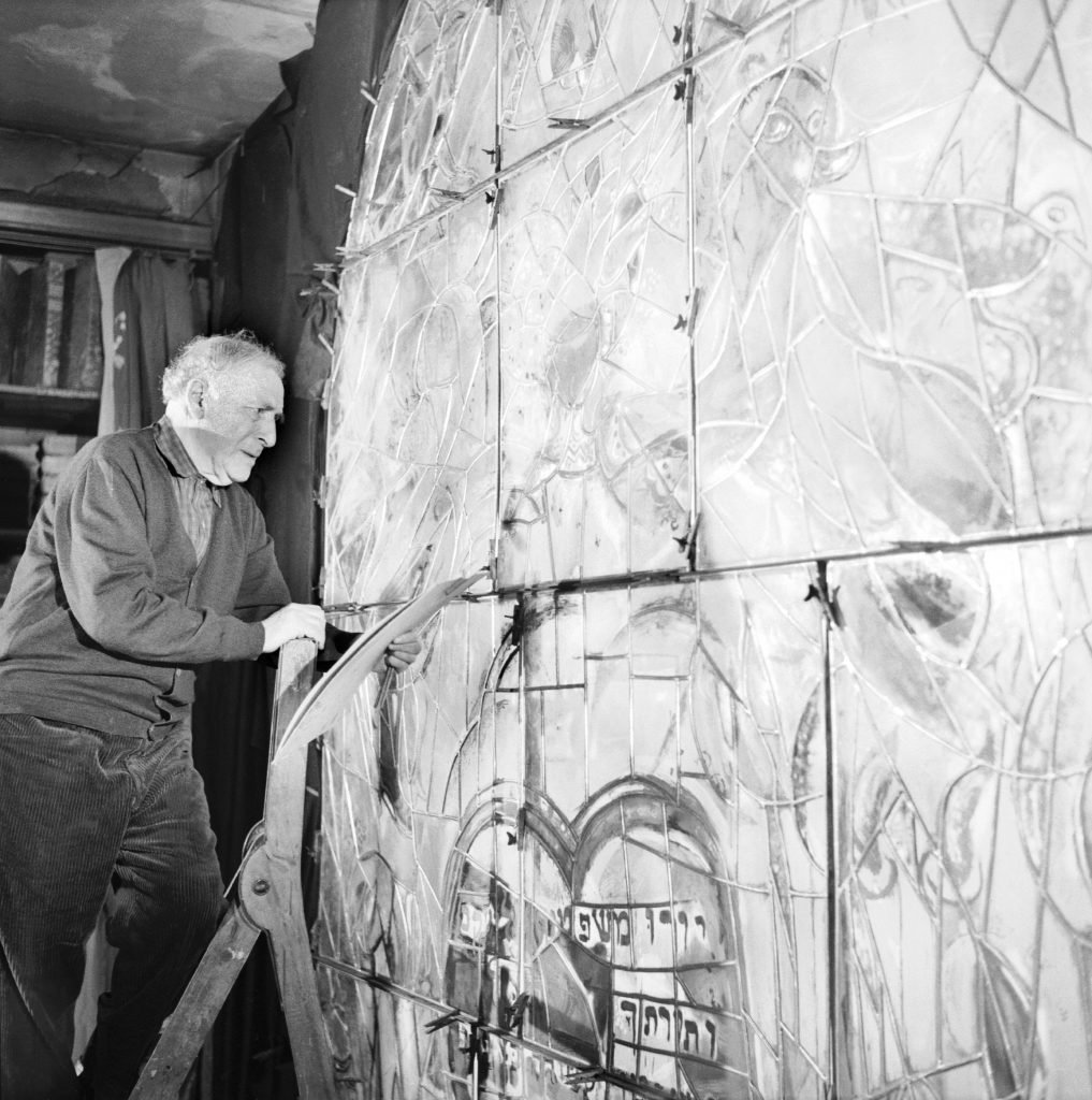 An elderly man standing on a ladder in front of panels of stained-glass, while holding a drawing board in his hand