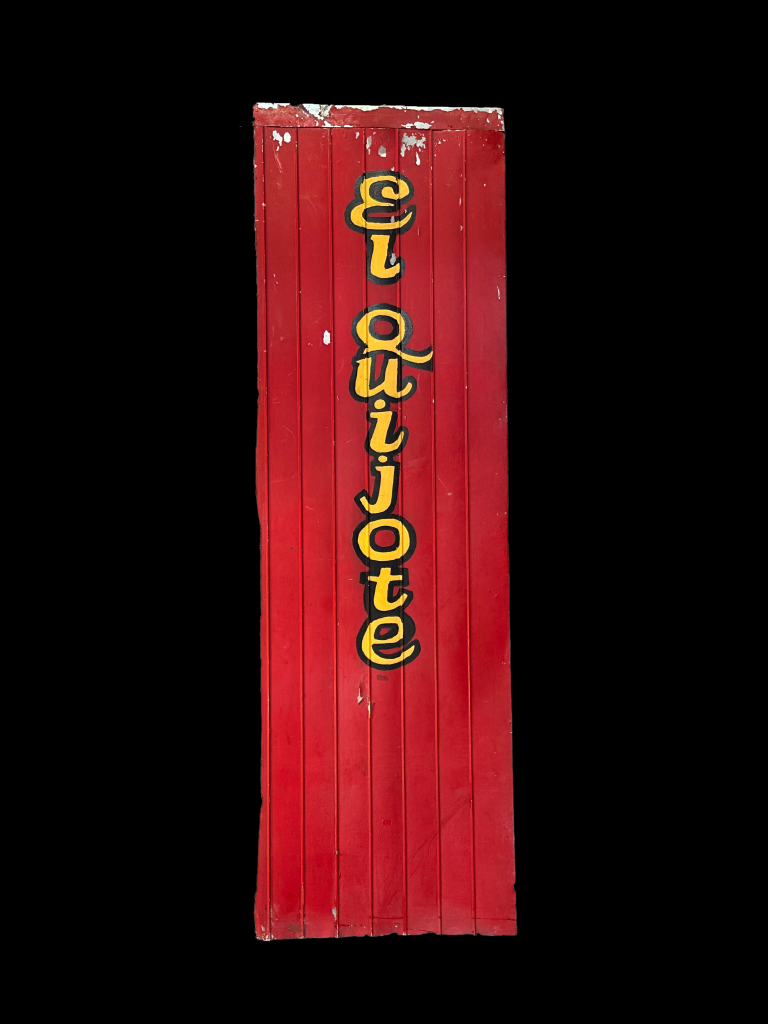 A photo of a tall red sign reading "El Quijoite" in yellow script with black outlines, on an all-black background.