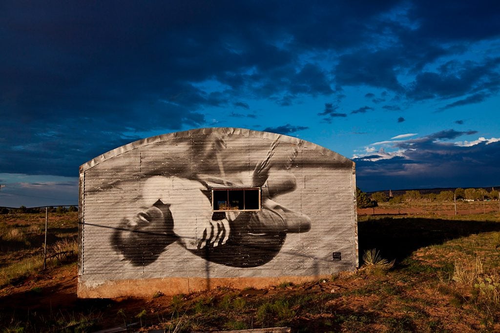 Featured in the 2024 FotoFocus Biennial, a photo of a side of a barn with a black and white photo of a person on a swing projected on it, and the sky is an oversaturated dark blue and scrub brush seen around it.