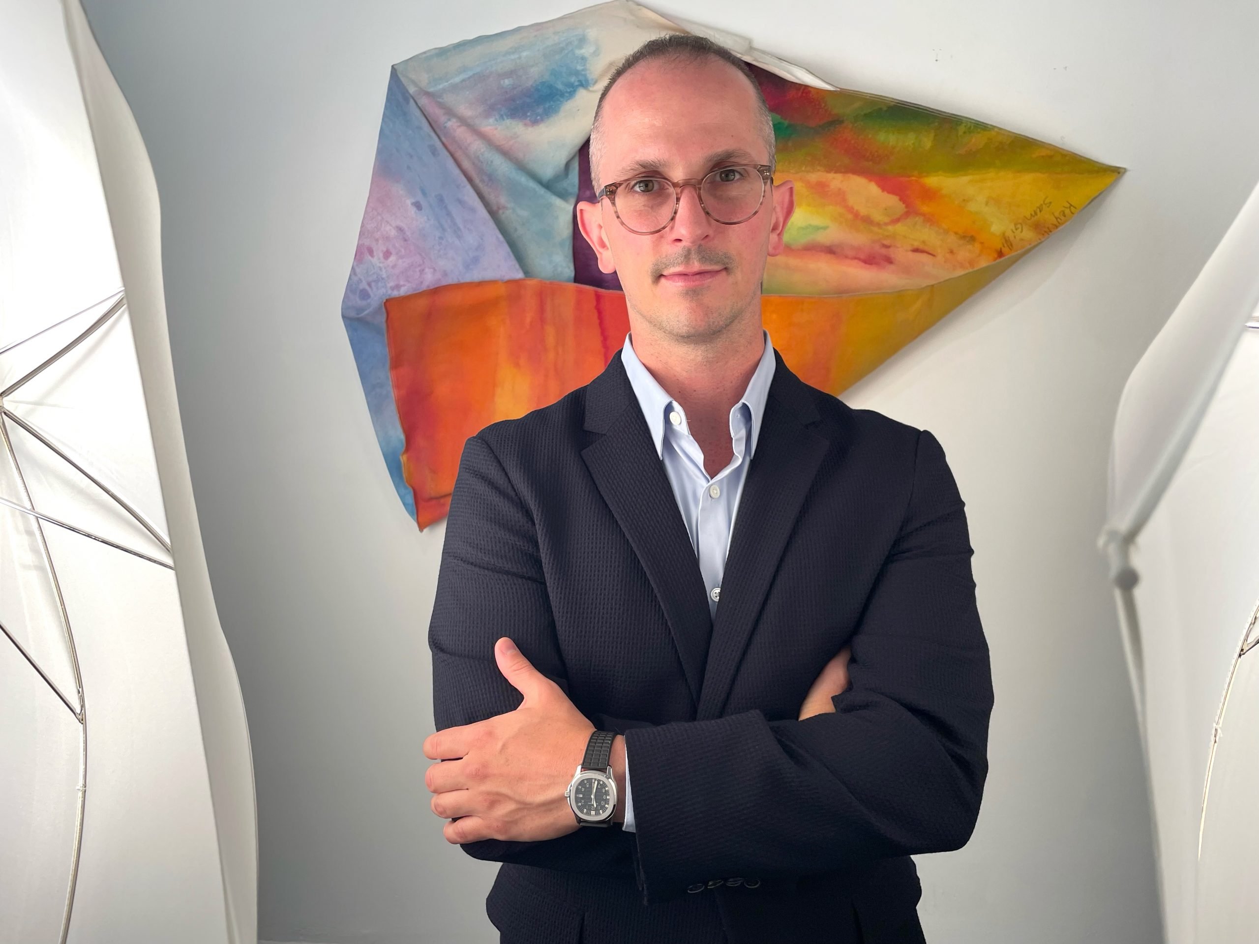 Half portrait of Dakota Sica wearing a suite and glasses with his arms crossed standing in front of an irregularly shaped abstract artwork in a white gallery space.