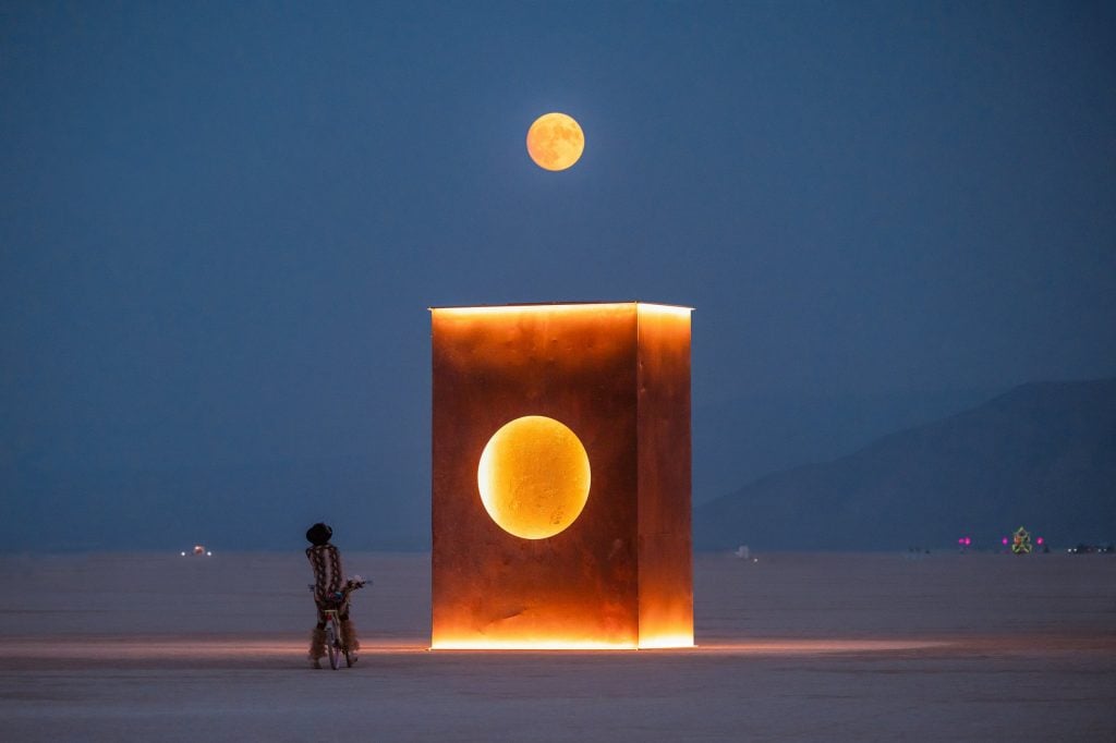 The image depicts a glowing rectangular sculpture by David Aiu Servan-Schreiber at the Burning Man gathering in the Black Rock Desert. The sculpture is a large, monolithic structure with a circular cutout in the center, illuminated with a warm, orange-yellow light. The full moon is positioned directly above the structure, aligning with the circular cutout, creating a surreal and harmonious visual effect. In the foreground, a person on a bicycle, dressed in a unique outfit, gazes at the installation, with the vast desert and distant mountains under the deep blue night sky.