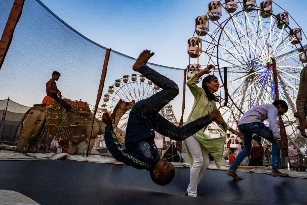 Low view photo of a family on a netted trampoline with a young boy upside down in the midst of a backflip. Two ferris wheels are visible behind.