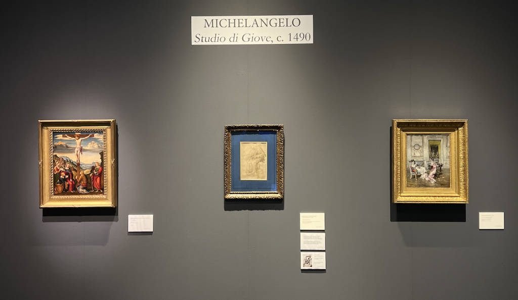 A photograph of three small framed old master drawings, included the Michelangelo drawing in the center, all mounted on a navy blue wall with a title plaque above them and wall text to their lower right hand corners.