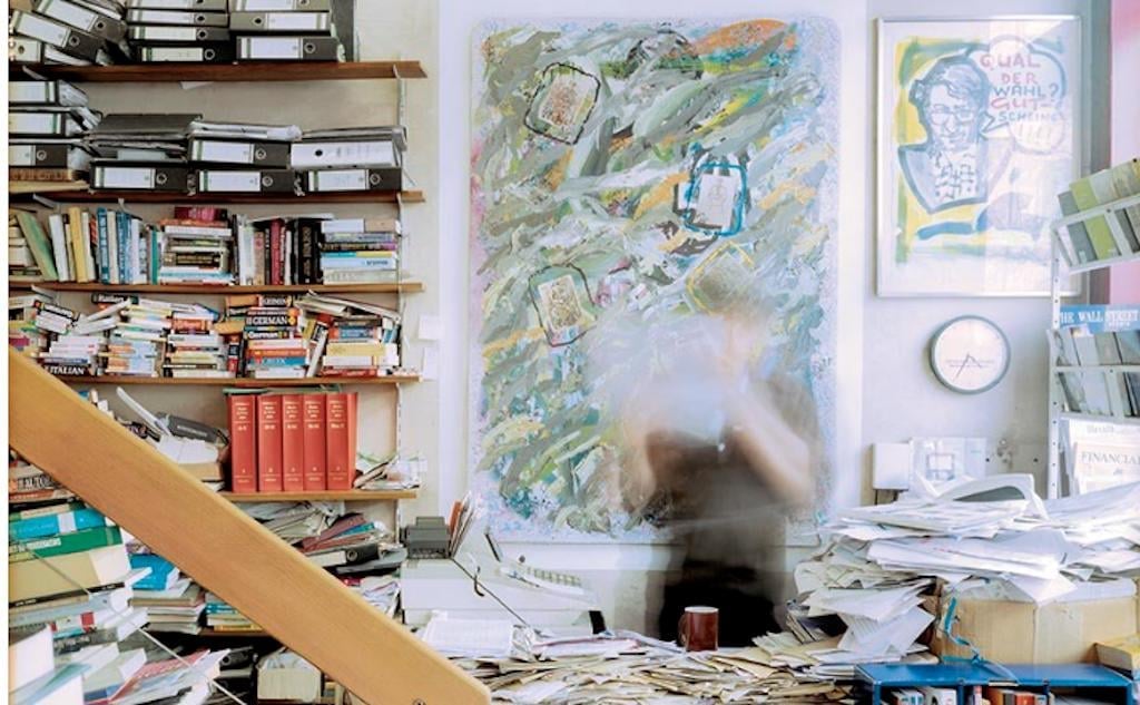 From an exhibition to see this month, a photo by dirk seiden schwan showing a cluttered office room with someone standing beind the desk but they are very blurry where everything else is clear.