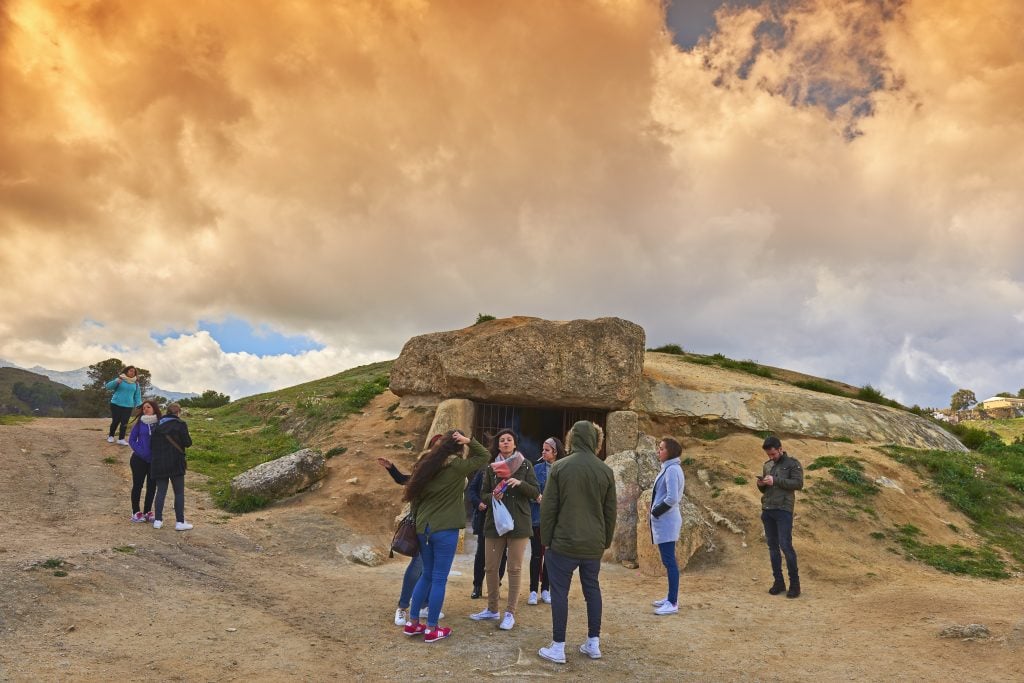 A photograph of a group of tourists standing in front of an ancient hillside burial place built from stones, atop dusty brown ground and beneath a cloudy sunset sky.