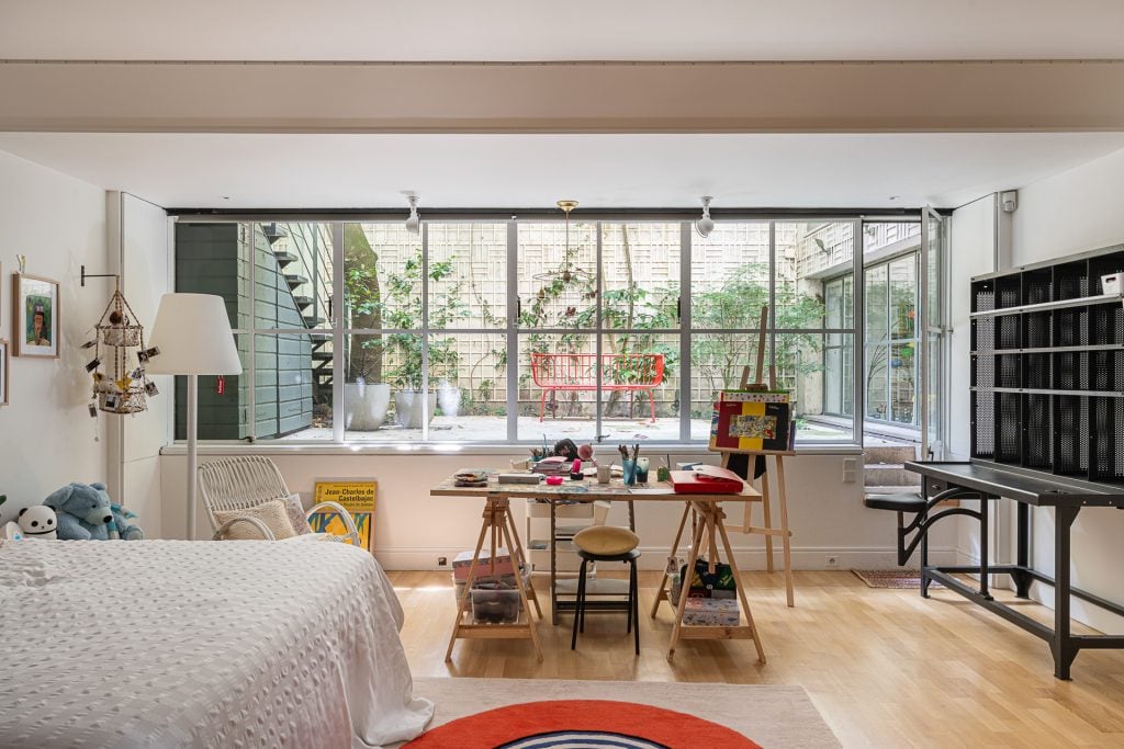 A bedroom with a cluttered desk and stuffed animals, anchored by a large window showing a terrace with a red bench