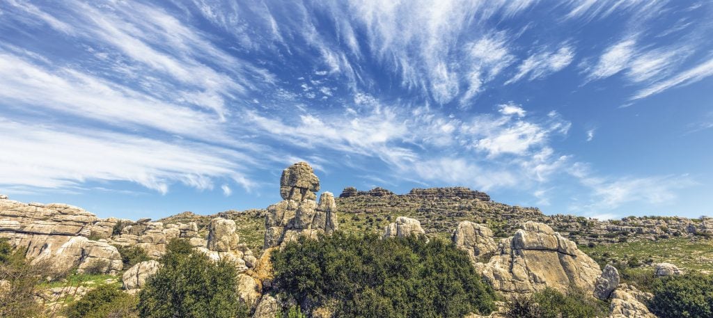 A photograph of a rock formation in Málaga, Spain speckled with sparse greenery beneath a blue sky with wispy white clouds spread throughout it.