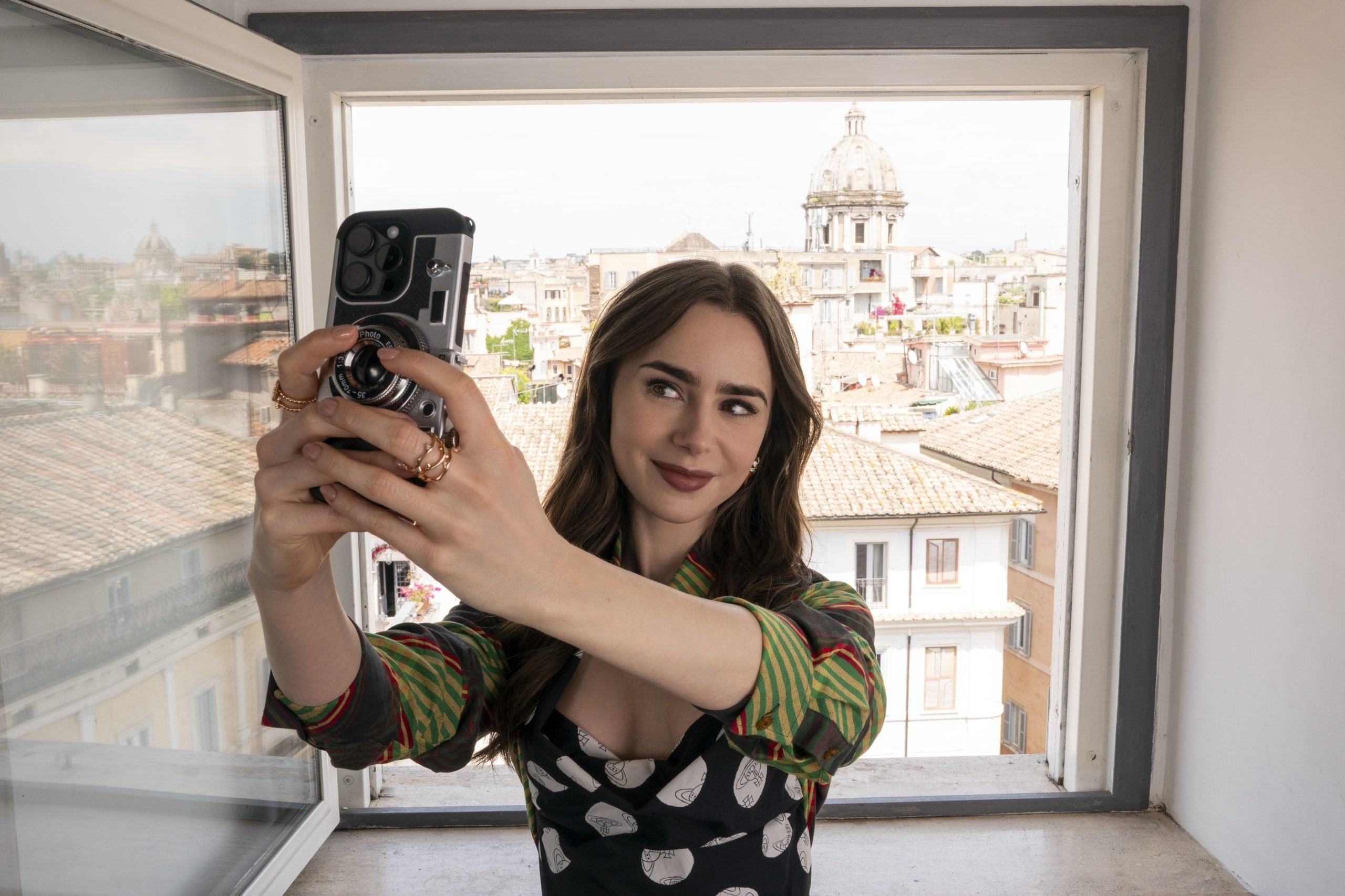 Lily Collins as Emily in Emily in Paris, season four, episode 10, taking a selfie out the window of her new temporary apartment in Rome. She has fair skin and long dark hair, wearing a dress with green and yellow striped sleeves and a black bodice with large white polka dots.