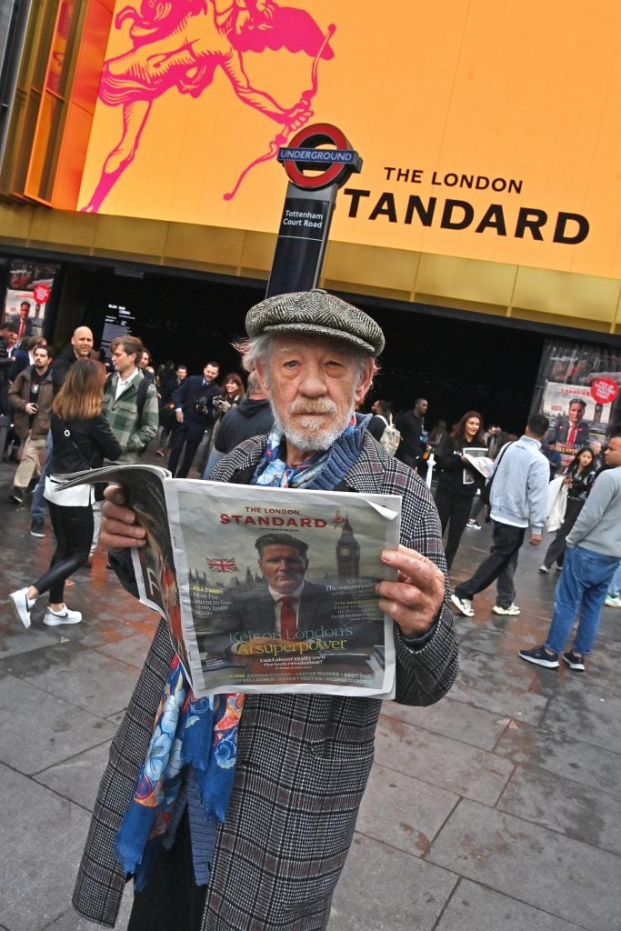 A man in a pageboy hat reads a tabloid newspaper on the street