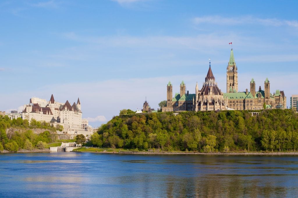 A scenic photograph depicting two majestic buildings peeking above trees and looming over a river, beneath blue skies.