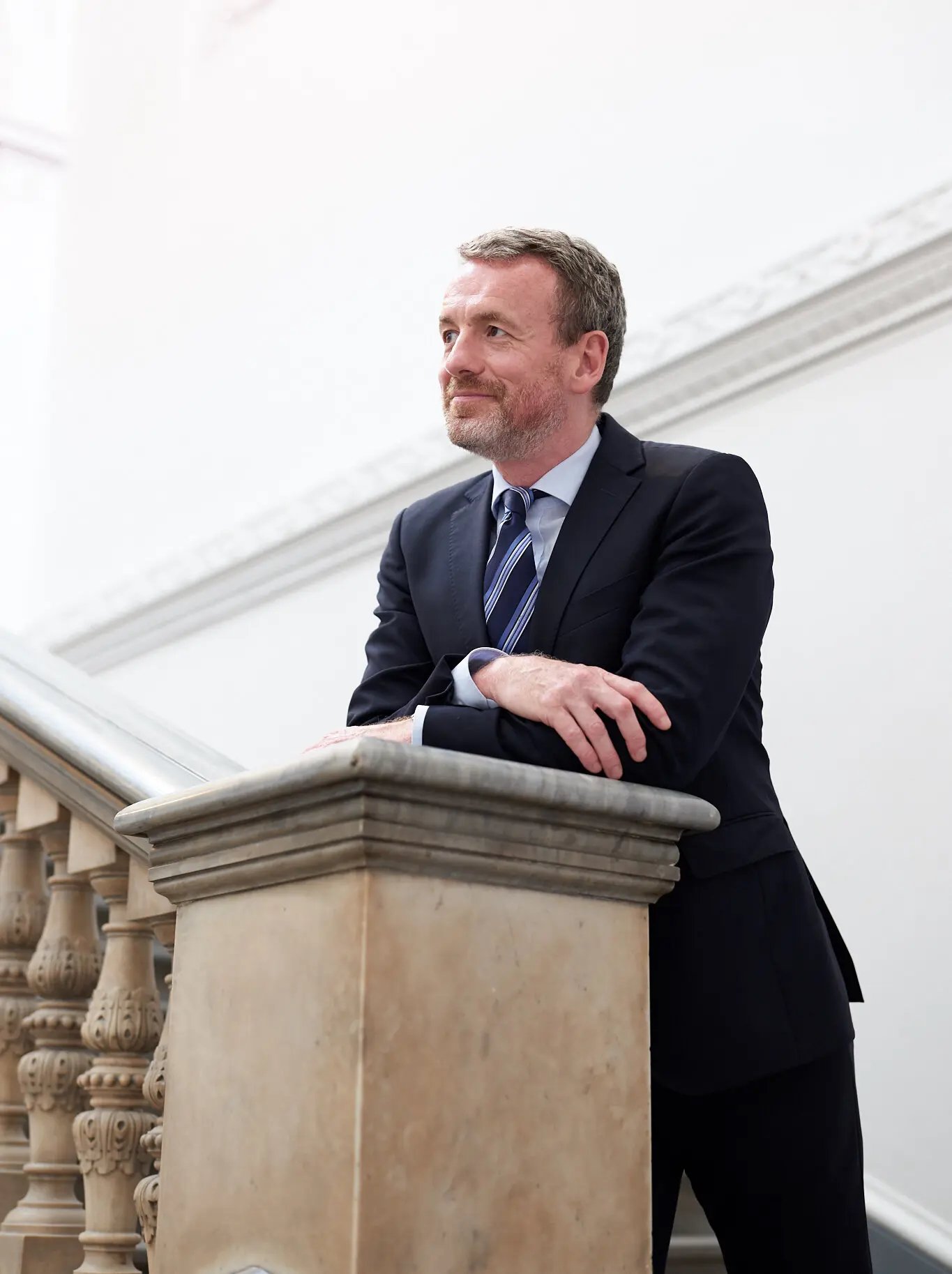 Axel Rüger, next leader of the Frick Collection, New York. A middle aged white man in a suit and tie leans over the end of an ornate stone bannister.