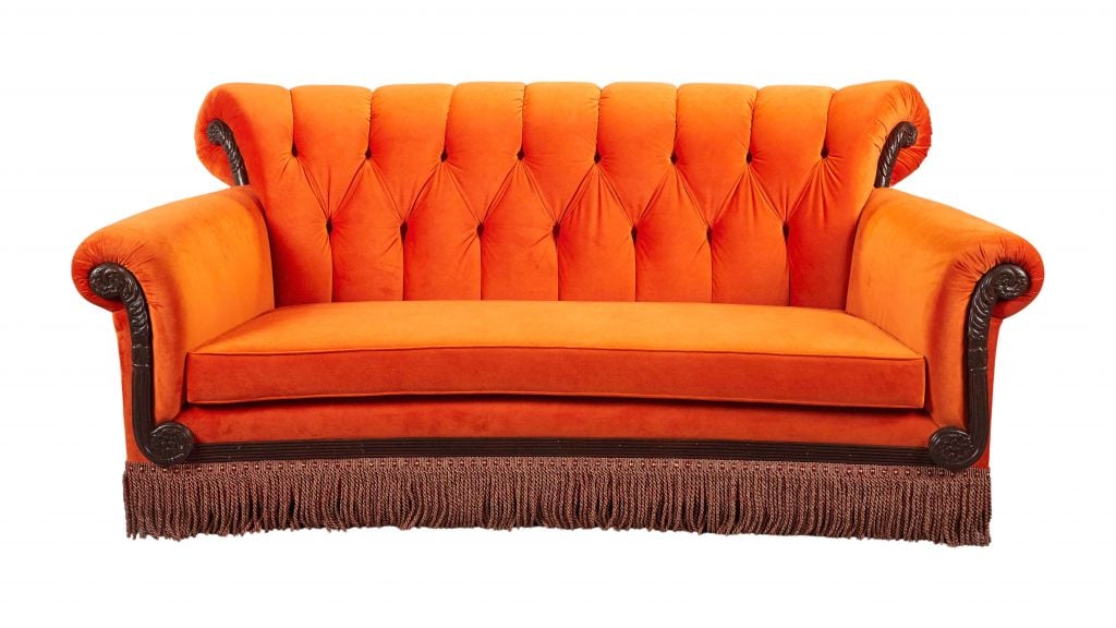 A photograph of the bright orange velvet couch from Friends depicted on an all white background.