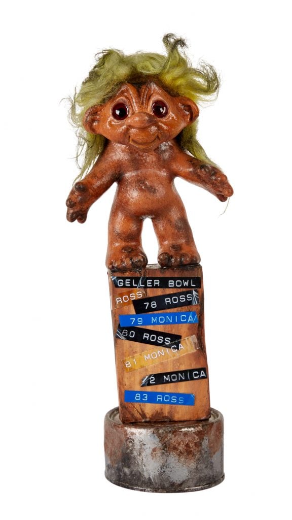 A photo of a makeshift trophy from the show Friends featuring a tattered Troll doll atop a wooden plank on a circular tarnished metal plinth, all depicted on an all-white background.