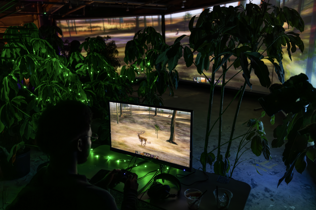 A photograph of a computer displaying a video game of a deer in a forest amongst an installation of potted plants, before a trifold of three larger screens displaying the same game.