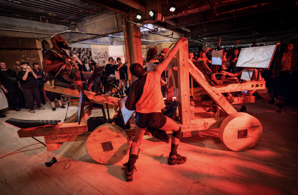 A photograph of performers acting atop a wooden catapault speckled with gaming screens, surrounded by a crowd and doused in orange light.