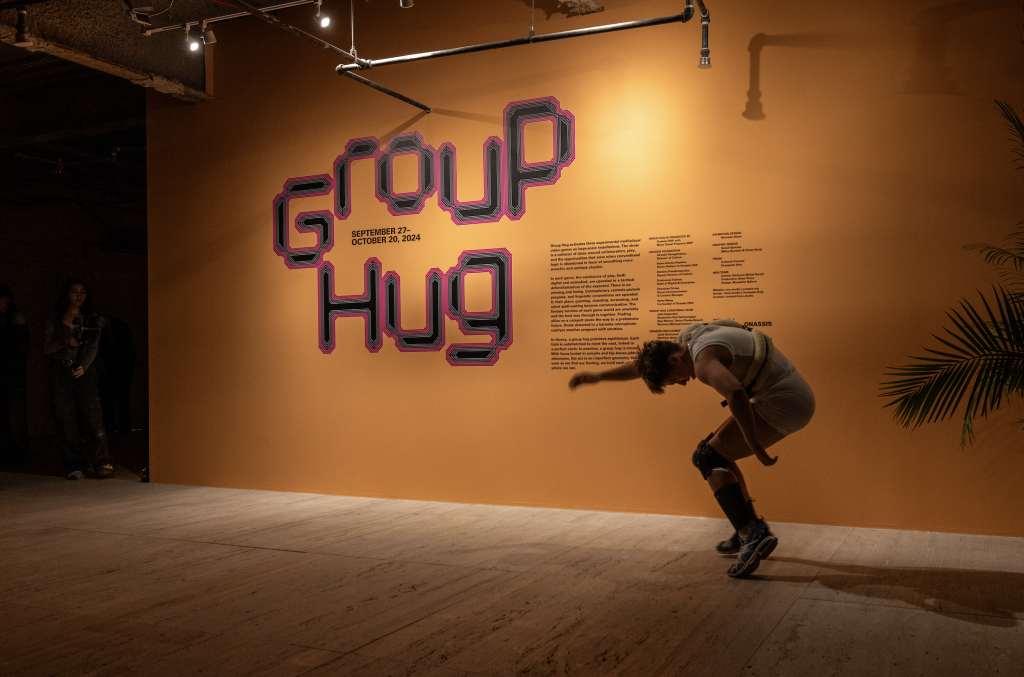 A photograph of a performer dancing before an orange wall labeled "Group Hug" with accompanying exhibiion introductory text.