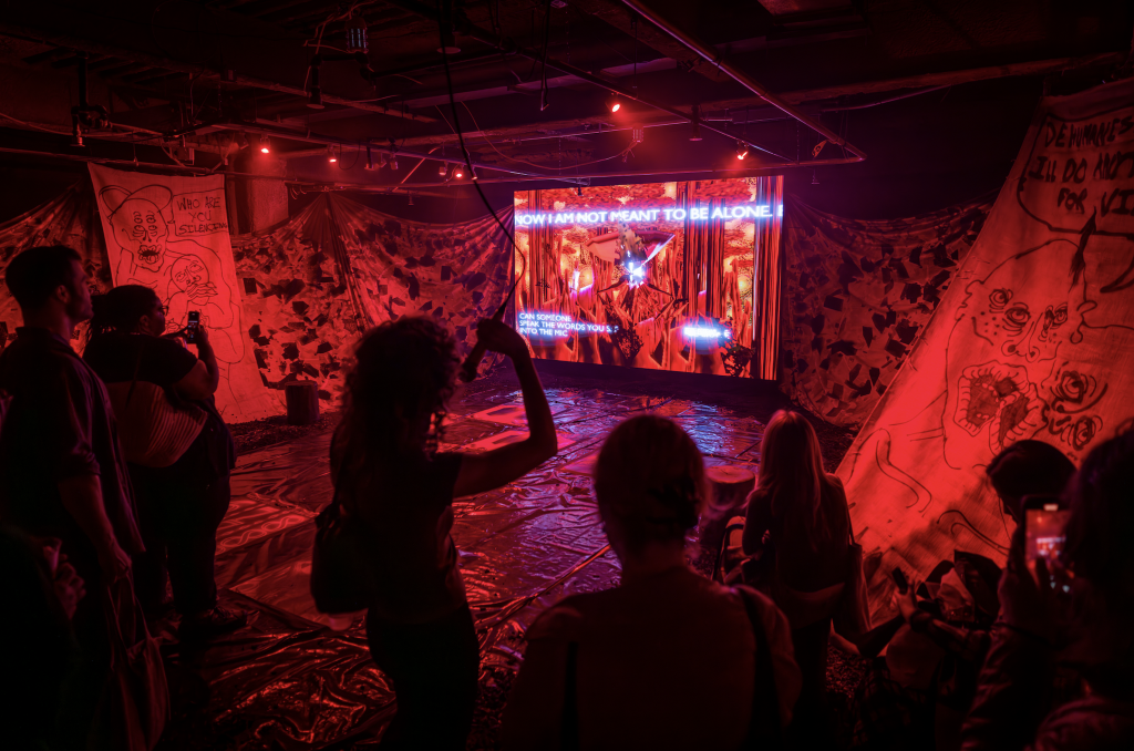 A photograph of a guest speaking into a hanging microphone in a red room where a video game is projected on a screen.