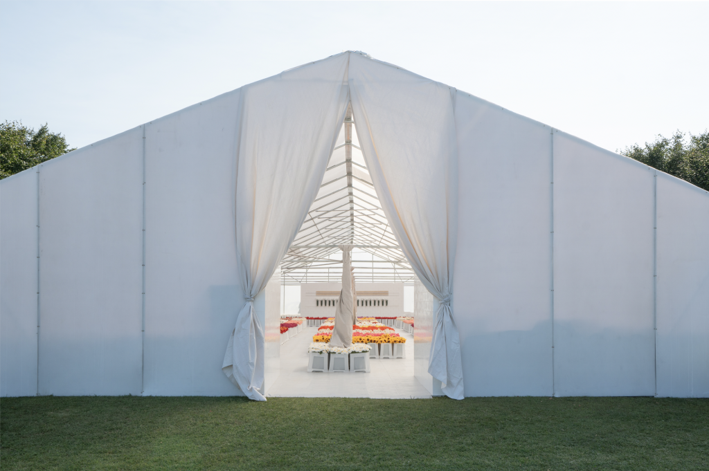 The exterior of a big white temporart tent with bis of flowers inside, atop green grass and beneath a clear sky.