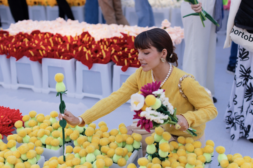 A photo of a girl wearing yellow kneeling down to pick yellow plush flowers from bins, with red plush lilies in the white bins behind her.