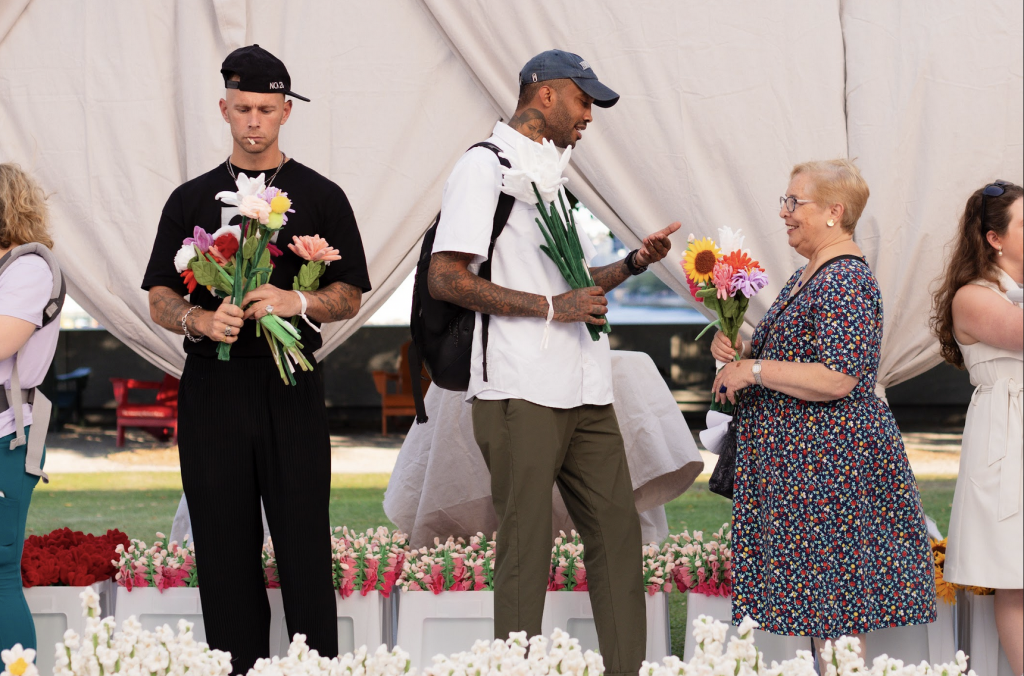 Three people hold plush bouqets beneath the white tent of Cj Hendry's "Flower Market"