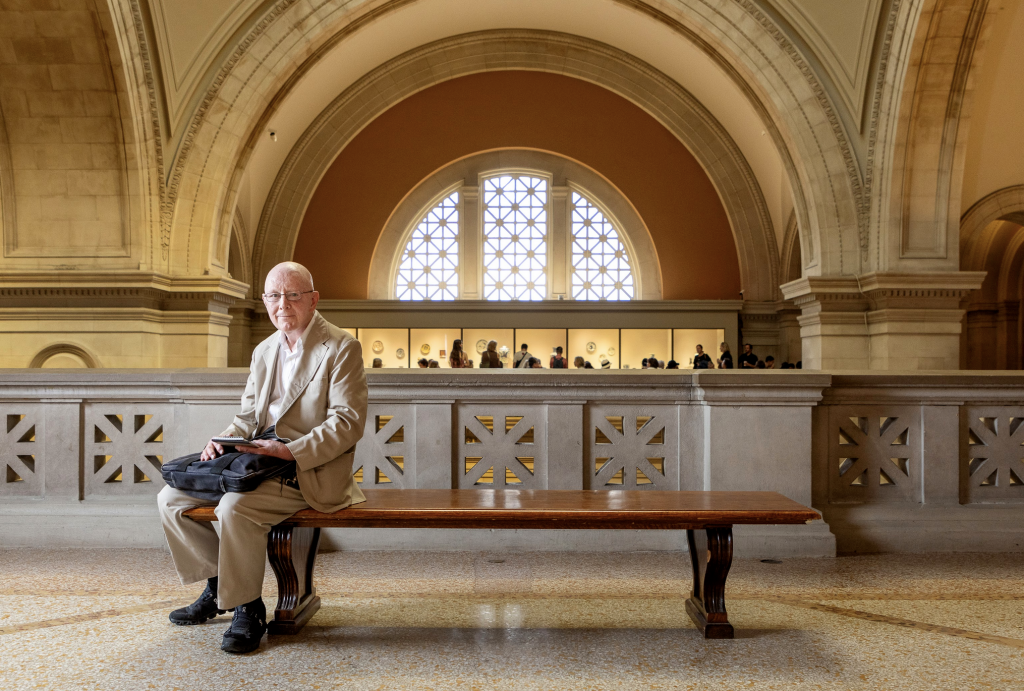 A photo of Holland Cotter dressed in a beige suit and seated on a bench amongst the marble mezzanine of an art museum, with an arched window behind him.