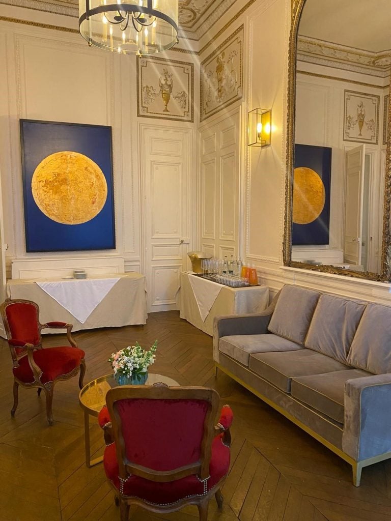 The image shows a refined meeting room at the Hotel Alfred Sommier in Paris, featuring David Aiu Servan-Schreiber's painting Coral Moon. The artwork, a vibrant depiction of a golden-yellow moon against a deep blue background, hangs on an elegantly paneled wall. The room is tastefully decorated with ornate moldings and a large gilded mirror reflecting the opposite side. In the foreground, two plush red velvet chairs surround a small coffee table with a vase of flowers, while a gray velvet sofa adds to the room's sophisticated ambiance. The parquet floor and soft lighting complete the luxurious setting.