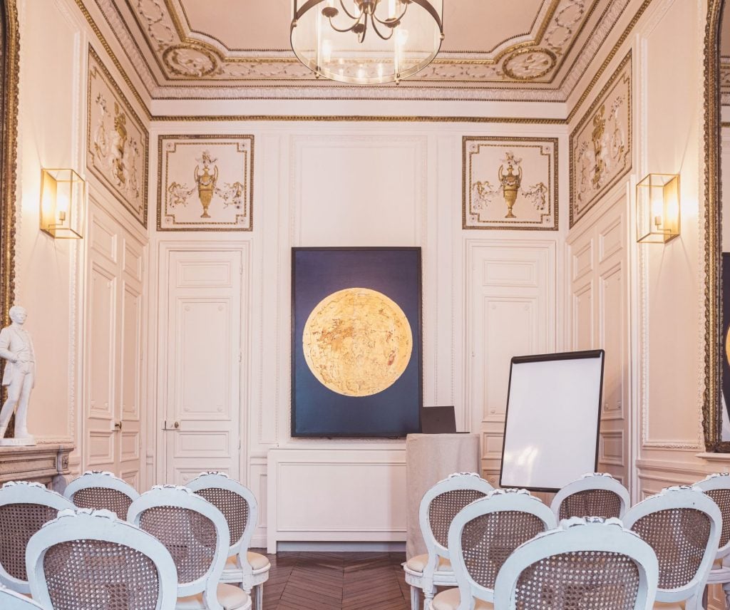 The image captures a luxurious meeting room at the Hotel Alfred Sommier in Paris, featuring David Aiu Servan-Schreiber's painting Coral Moon. The artwork, a large golden moon set against a deep blue background, hangs prominently on the intricately decorated wall. The room is adorned with classical French design elements, including ornate moldings, a chandelier, and gilded details on the ceiling. In front of the painting, a small podium and a blank presentation board are set up, while several elegant white chairs with woven backs are arranged for a meeting or event. A white statue stands to the side, adding a touch of classicism to the refined atmosphere.