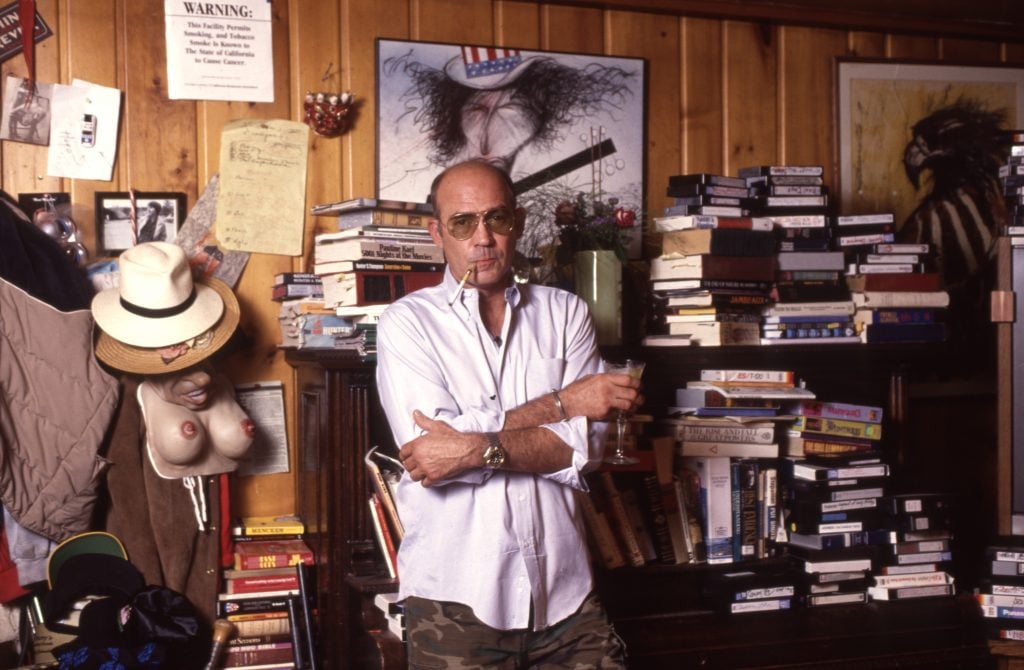 Journalist Hunter S. Thompson in sunglasses standing in a wood-paneled room filled with books and a picture by Ralph Steadman