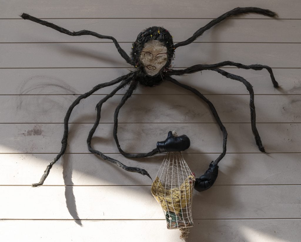 A spider sculpture with a human face and two gloves on two of its eight legs holding a mesh bag with a rooster object.