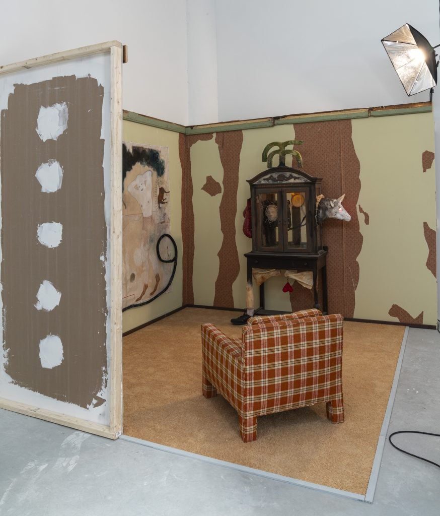 Installation view of Amy Bravo solo show where a sitting room has been recreated with a plaid armchair, peeling wallpaper and a curio cabinet sculpture.