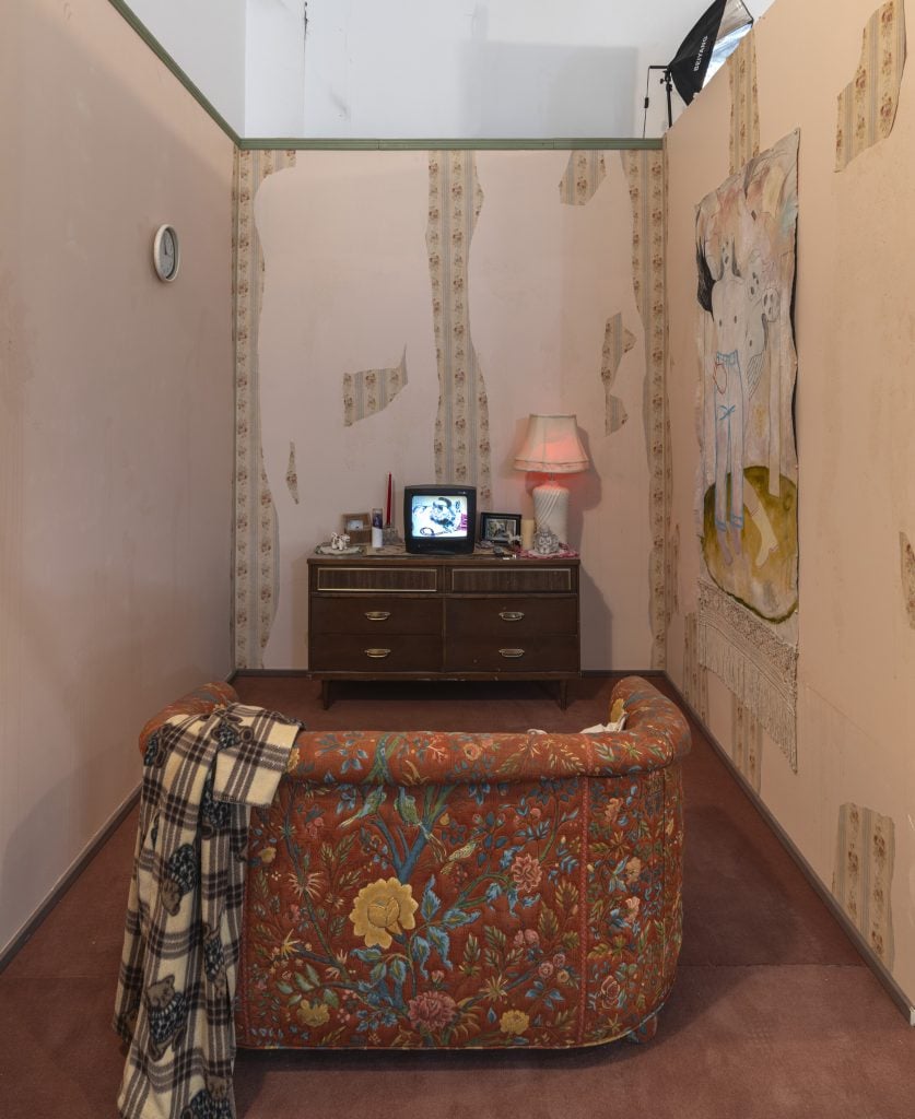 Inside a room made to look like a grandma's living room, with a small patterned loveseat, small tube television, a table lamp with pink lampshade, and peeling floral wallpaper. Inside Amy Bravo solo exhibition at Swivel Gallery.