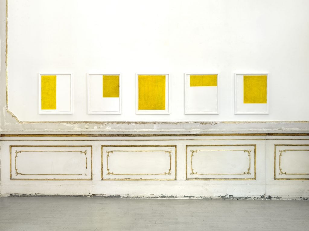 View of a wall with aged Rococo style panelling along within are five small-scale paintings by Callum Innes with one different size and placed yellow rectangle on each.