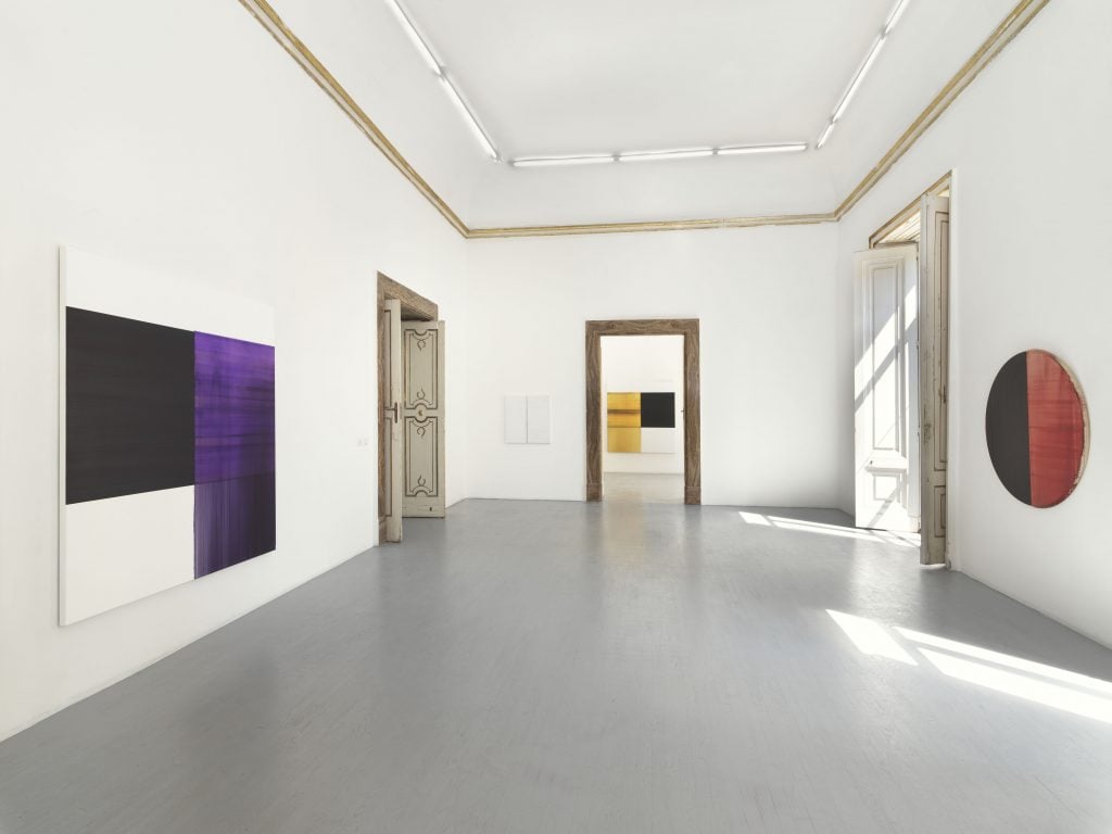 Inside Alfonso Artiaco, Naples, gallery space with a door length window on the right letting in light, a doorway on the far wall through which you can see a painting by Callum Innes and on the right wall a tondo painting half black half red, and on the left wall another doorway and a square painting painted in blacks and purples in squared composition.