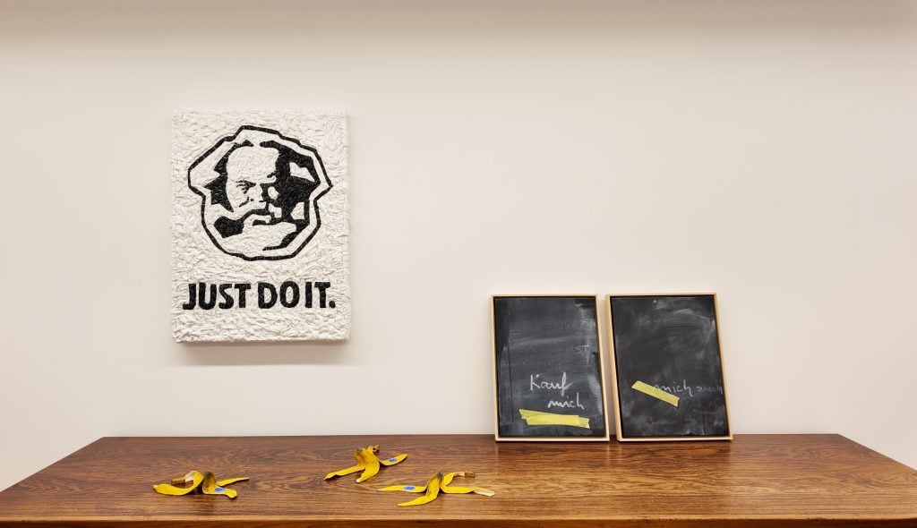 Installation view of exhibition at Filser and Gräf showing the work of three artists next to each other, three banana peel sculptures by Jessi Strixner on top of a bench, a white composition made out of garbage bags showing a portrait of Karl Marx with the slogan "Just do it" underneath, and two acrylic paintings leaning against the wall.