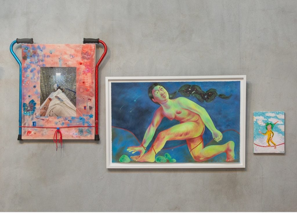 Three paintings on a gray wall, on the left the canvas as a colorful contemporary cane on the right and left, the center painting a figurative work of a woman lunging forward and looking back against a dark blue sky, and on the right a diminutive abstract work.