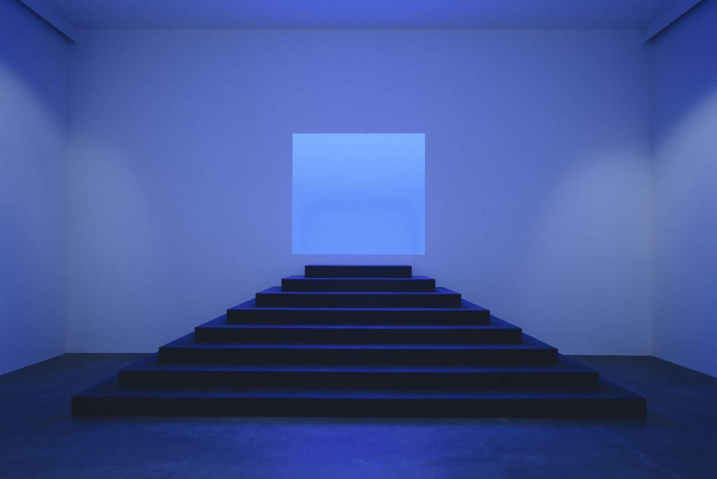 A photograph of an immersive, blue-tinged installation by James Turrell where dark steps lead to a luminous square