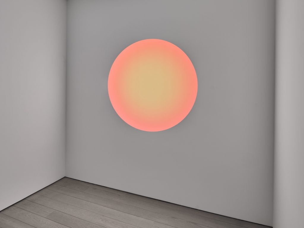 A photograph of a glowing orange circular led artwork on a white wall before a white floor.