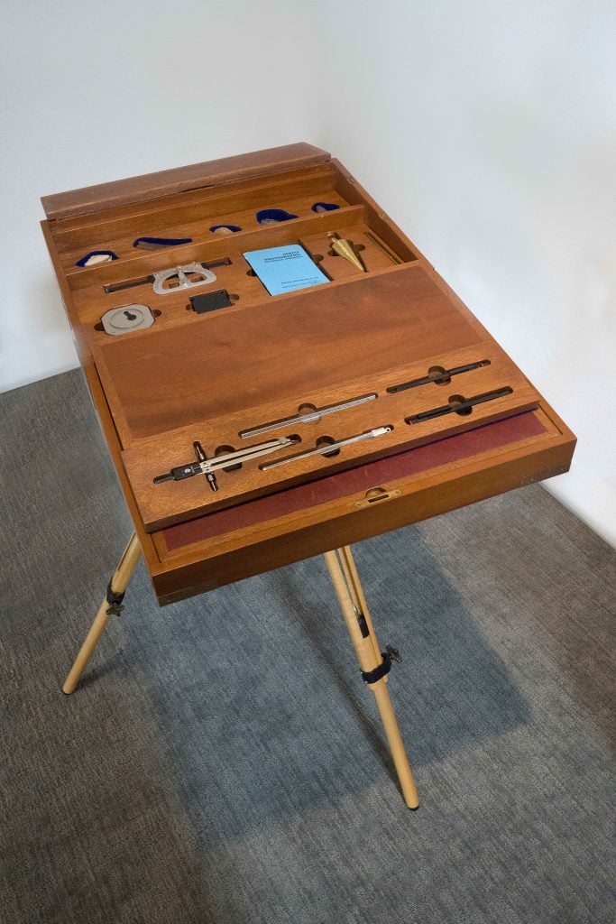 A photo looking down on a wooden lap desk with numerous compartments made for differently shaped drafting tools, standing on a gray floor surrounded by white walls.