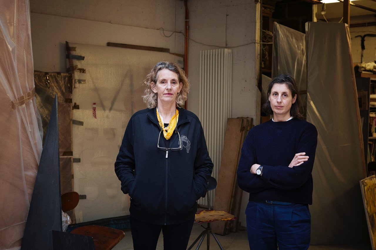 Portrait of the artist duo Cullinan Richards showing two women, Jeanine Richards and Charlotte Cullinan, standing next to each other in a shadowy studio room. Both are wearing black.