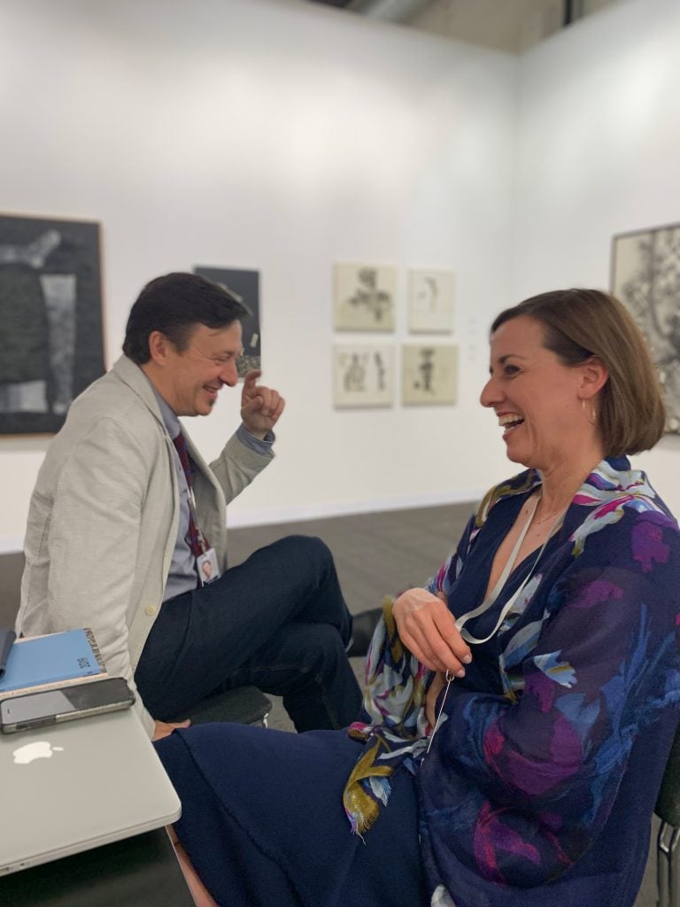 A seated man and woman laugh, with artworks behind them.